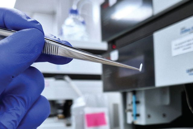 Gloved hand holding a solid-dose vaccine pellet in tweezers in a laboratory